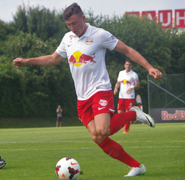 Ein professioneller Fussballspieler in einem roten Trikot kickt einen Ball auf einem grünen Feld mit Bäumen und einem klaren blauen Himmel im Hintergrund, in der Nähe einer Tafel mit der Aufschrift "RB Leipzig."
