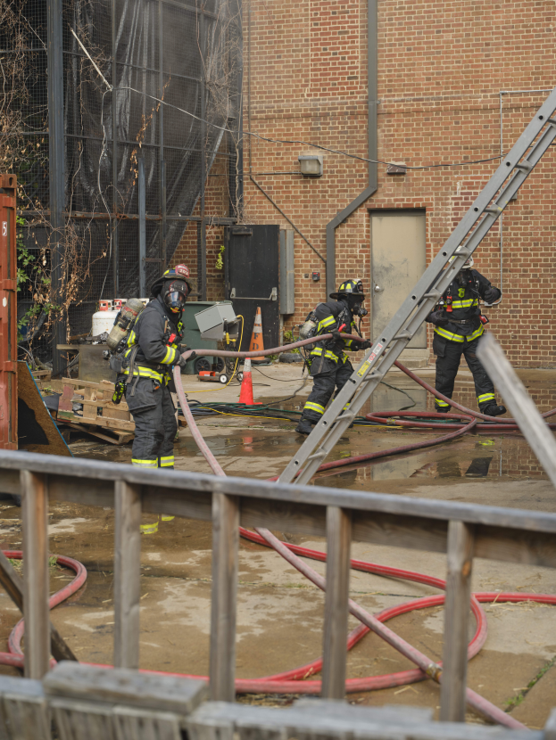 Eine Gruppe von Feuerwehrmännern in Helmen, die Rohre halten, arbeiten daran, ein Gebäude Feuer zu löschen, mit verschiedenen Geräten und Strukturen drumherum, einschließlich eines Metallzauns, Rohre auf dem Boden, einem Container, einem Verkehrskegel und einem Baum im Hintergrund.