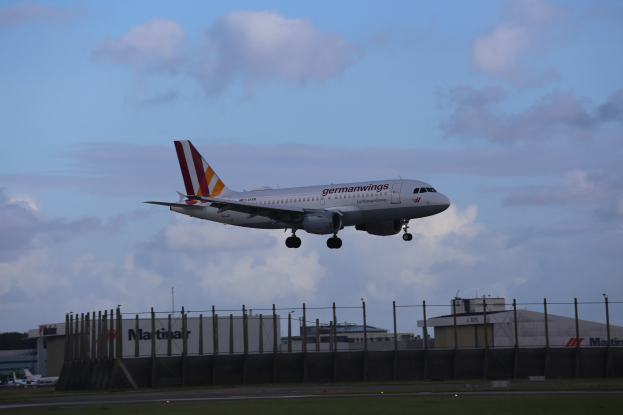 Germanwings Airbus A320-200 beim Abheben vom Frankfurter Flughafen, mit einem Zaun im Vordergrund, Gras darunter, Gebäuden und Bäumen im Hintergrund und einem bewölkten Himmel darüber.