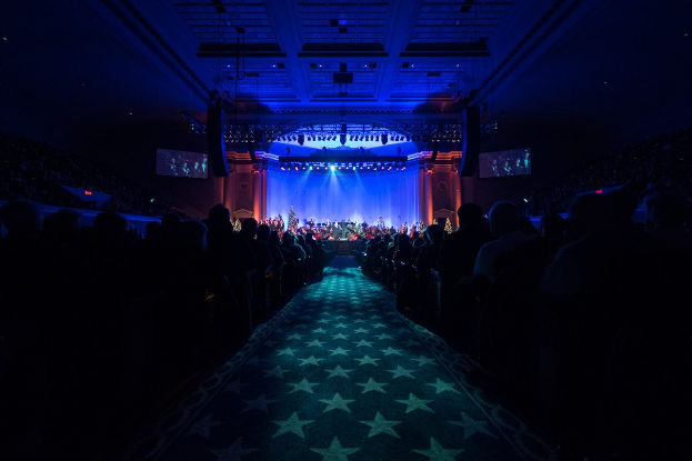Großer Saal mit sitzendem Publikum, Bühne mit Musikern und Deckenleuchten und -fernsehern in einer dunklen Konzerthalle.