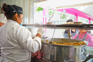 Eine Frau in Kochuniform, weißem Kleid, Handschuhen und schwarzer Mütze, hält einen Löffel und steht vor einer Stahlschüssel voller Essen, mit einer anderen Frau in blauem Kleid links von ihr und diversen Küchenutensilien und Menschen im Hintergrund.