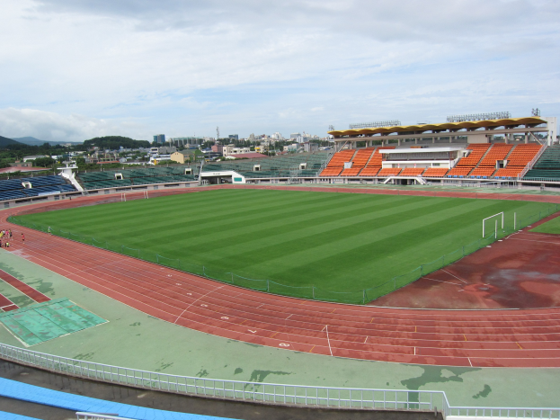 Großes Stadion mit einem Fußballfeld, umgeben von Gebäuden, Bäumen, Hügeln und einem klaren blauen Himmel, mit wenigen Zuschauern auf saftig grünem Gras.