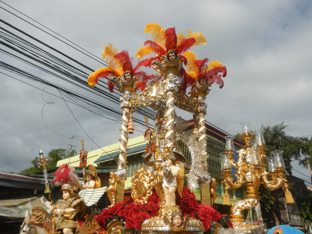 Ein geschmückter Rosenmontagszug mit Statuen und Blumen, umgeben von Gebäuden, Bäumen, Polen, Drähten und dem Himmel oben.