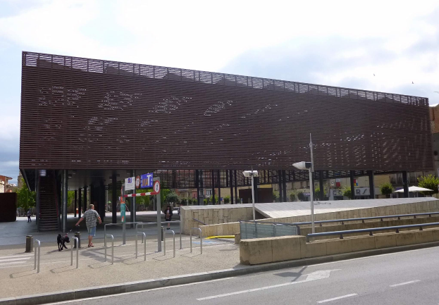 Großes modernes Gebäude mit zahlreichen Fenstern, das als neuer Eingang zum Stadtzentrum dient, umgeben von städtischen Elementen wie Straßenlaternen, Fahrzeugen, Fußgängern und Bäumen unter einem bewölkten Himmel.