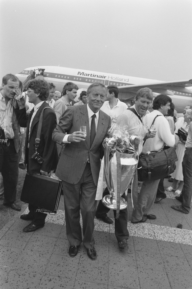 Ein Mann im Anzug und Krawatte hält eine Trophäe vor einem Flugzeug, umgeben von einer Gruppe von Menschen, einige halten Taschen und einer hält eine Kamera, in einem Schwarz-Weiß-Bild.