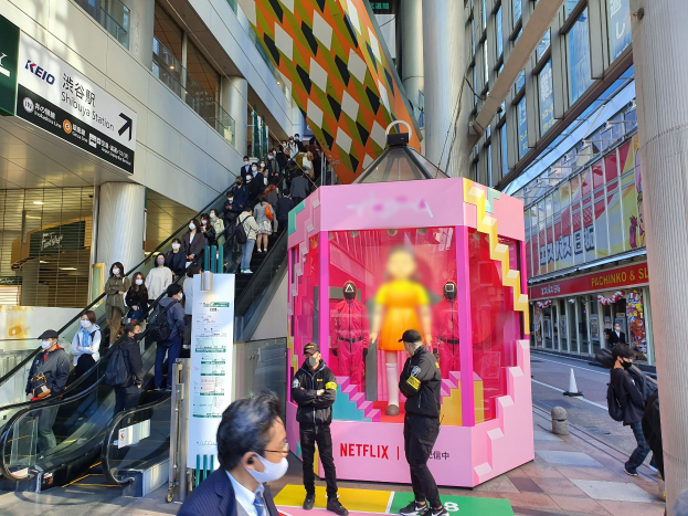 Eine Gruppe von Menschen vor einem Glaswandgebäude mit Texttafeln, einige tragen Masken, in der Nähe einer Rolltreppe; eine pinkfarbene Box in der Mitte zeigt "Netflix's new Netflix-Booth in Tokyo" mit Schaufensterpuppen darin.