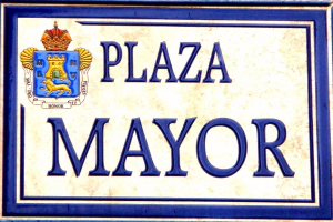 Rechteckiges weißes Schild mit schwarzer Schrift "Plaza Mayor" in fetter Schrift, blauem Rand und einem gekrönten Wappen-Logo an einer Gebäudefassade.
