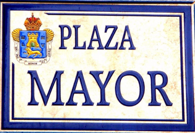 Rechteckiges weißes Schild mit schwarzer Schrift "Plaza Mayor" in fetter Schrift, blauem Rand und einem gekrönten Wappen-Logo an einer Gebäudefassade.