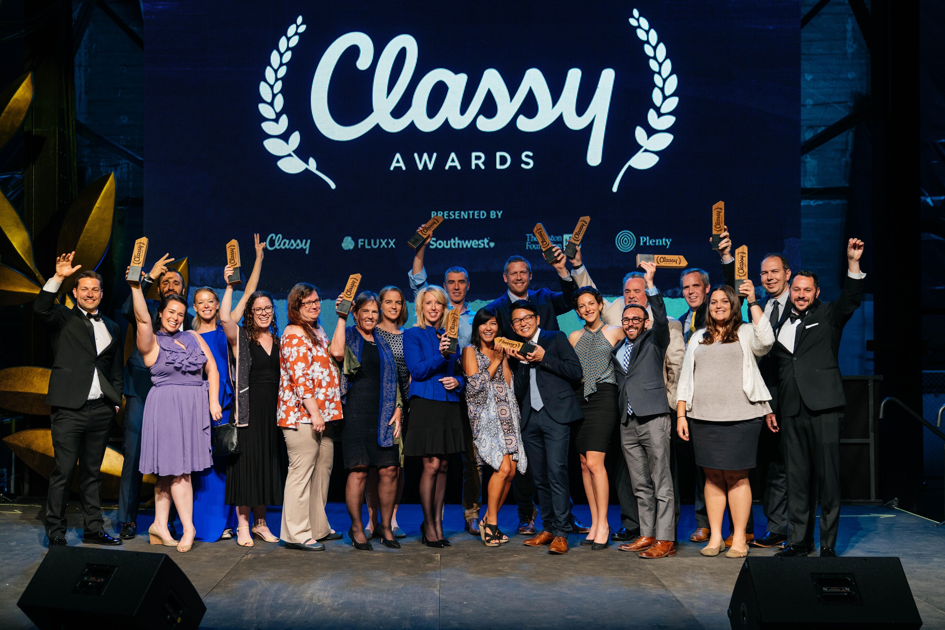 Gruppe von Menschen auf einer Bühne mit Auszeichnungen, wobei "Classy Awards 2019" auf einem Bildschirm dahinter angezeigt wird.
