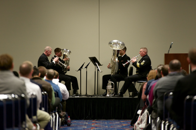 Eine Gruppe von Männern, die auf einer Bühne Blasinstrumente spielen, mit Notenständern, einem Podium mit Mikrofon und einer Sitzreihe mit Zuschauern im Hintergrund.