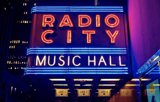 Radio City Music Hall in New York City, hell erleuchtet und von hohen Gebäuden flankiert, mit einer "Radio City"-Leuchtschrift und zusätzlichen Schildern auf der rechten Seite.