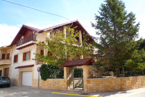 Eine Villa mit einem davor geparkten Auto, das Fenster, Geländer, Säulen und ein Dach hat, umgeben von einem Tor, einem Schild, einem Straßenpfahl, Bäumen und einer Wand unter einem bewölkten Himmel.