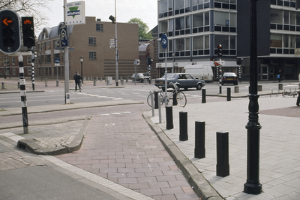Eine Straßenszene in der Stadt mit einem geparkten Fahrrad, fahrenden Fahrzeugen, Fußgängern, Pfosten, Verkehrsampeln, Schildern, Bäumen, Gebäuden mit Fenstern und einem klaren blauen Himmel.