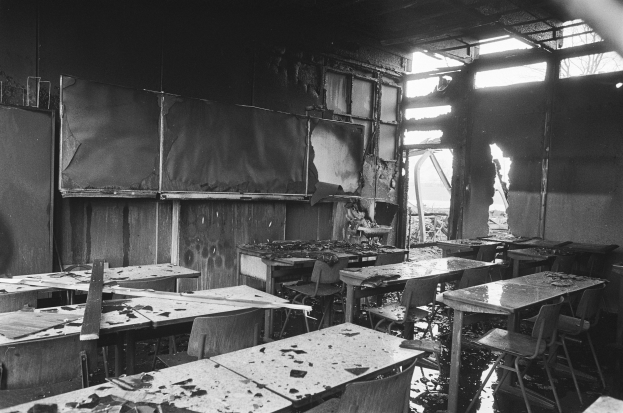Schwarz-weißes Foto eines verlassenen Klassenzimmers mit verkohlten Wänden, zerbrochenen Fenstern und verstreuten Tischen und Stühlen.