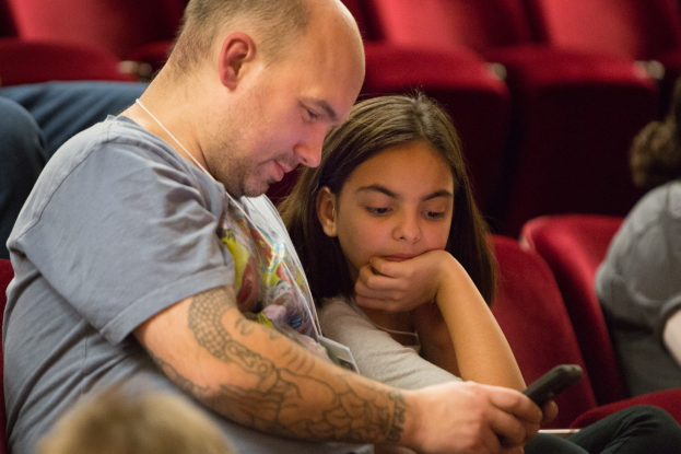 Zwei Menschen, ein Mann und ein junges Mädchen, sitzen in einem Kino und sind beide auf ein Handy konzentriert, das der Mann hält.