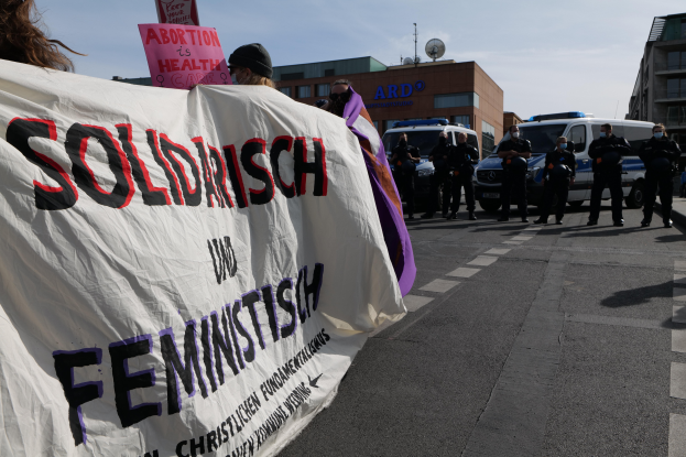 Eine Gruppe von Menschen marschiert mit einem Banner, auf dem "Solidarität und Feminismus" steht, eine Straße entlang, mit parkenden Fahrzeugen und Gebäuden im Hintergrund unter einem klaren blauen Himmel.