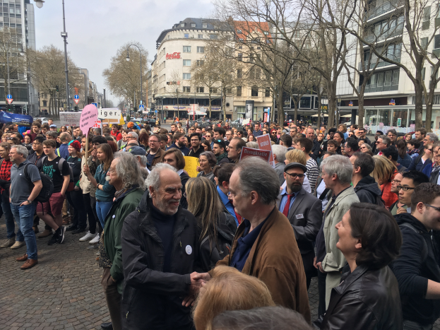 Eine große Gruppe von Menschen steht vor einer Menge mit Schildern, mit Fahrzeugen, Bäumen, Laternenpfählen und Gebäuden im Hintergrund, wahrscheinlich während einer Demonstration in Berlin, Deutschland.