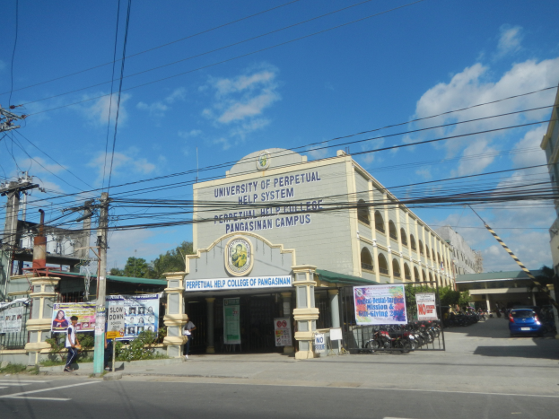 Die Universität Perpetual Help System in Pasig, mit Gebäuden, Fahrzeugen auf der Straße, Fußgängern, Werbetafeln, Strommasten, Kabeln, Bäumen und einer bewölkten Himmel.