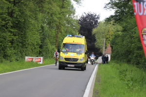Ambulanz fährt auf einer Straße mit Radfahrern nebenher, Gras und Bäume auf beiden Seiten, Häuser, Masten und einen klaren blauen Himmel im Hintergrund.
