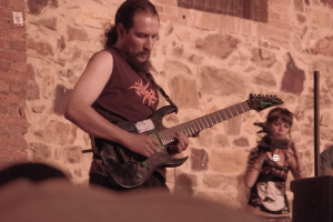 Ein bärtiger Mann spielt Gitarre auf der Bühne, rechts neben ihm hält eine Frau ein Mikrofon und im Hintergrund ist eine Menschenmenge zu sehen.