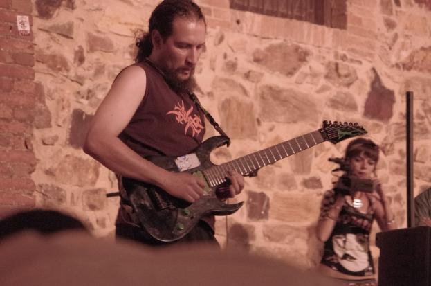 Ein bärtiger Mann spielt Gitarre auf der Bühne, rechts neben ihm hält eine Frau ein Mikrofon und im Hintergrund ist eine Menschenmenge zu sehen.