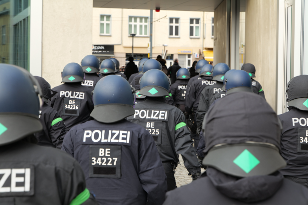 Eine Gruppe uniformierter Polizisten mit Helmen steht vor einem Gebäude, mit Menschen, Pfählen und anderen Gebäuden im Hintergrund.