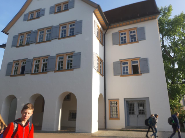 Eine Gruppe von Menschen, einige mit Taschen, geht vor einem großen weißen Gebäude mit Fenstern und Türen vorbei, das von Bäumen umgeben ist und unter einem klaren blauen Himmel steht, was darauf hindeutet, dass sie auf dem Weg zu einer Schule in Deutschland sind.
