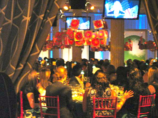 Eine Gruppe von Menschen, die an Tischen sitzen und Essen, Fernseher, Lampen, hängende Papiere und Vorhänge in einer Party-Atmosphäre sehen.