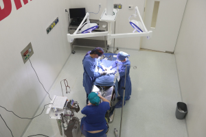 Chirurgen führen eine Operation an einem Patienten in einem Operationssaal durch, umgeben von medizinischer Ausrüstung und einer Wandtexttafel und einem Bodenobjekt im Hintergrund.