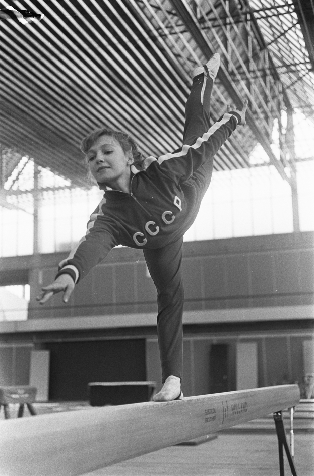 Ein junges Mädchen führt Gymnastik auf einem Balken in einer Turnhalle durch, mit einer Wand im Hintergrund und einer Decke mit Stangen oben.