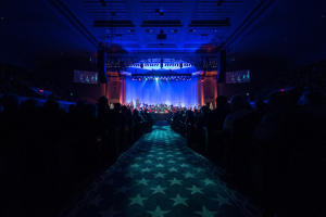 Ein großer Auditorium mit Sitzplätzen, eine Bühne mit Musikern und Deckenbeleuchtung und Bildschirme, was auf ein Konzerthaus während der Nationalkonvention 2019 hinweist.