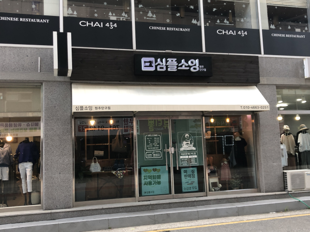 Außenansicht des Chai 434 Chinesischen Restaurants in Seoul, Südkorea, mit Glaswänden, Schaufensterpuppen in Kleidern, beleuchteten Schildern und einer Klimaanlage auf der rechten Seite, wobei unten eine Straße zu sehen ist.