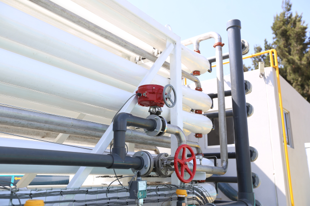 Große Gruppe von Rohren, Ventilen, Metallstangen und anderen industriellen Komponenten auf einem Gebäudedach mit Bäumen und einem klaren blauen Himmel im Hintergrund.