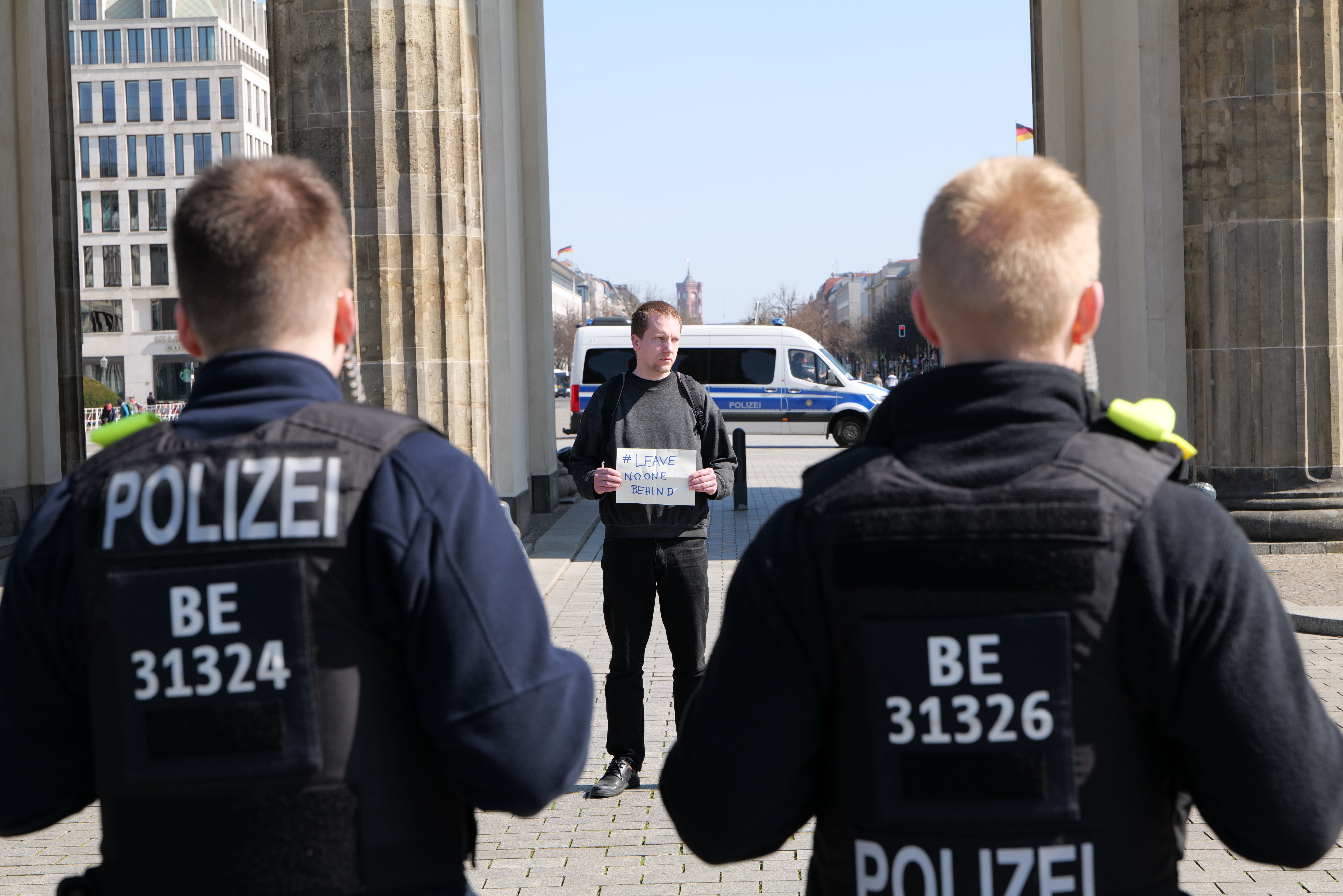 Drei Polizeibeamte stehen vor einem Gebäude, mit einem Mann in der Mitte, der ein Blatt hält, flankiert von Säulen, mit Fahrzeugen, Menschen, Bäumen, Gebäuden und dem Himmel im Hintergrund.