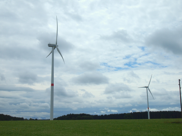 Drei hohe weiße Windräder in einer grünen Wiese mit Bäumen und Wolken im Hintergrund.