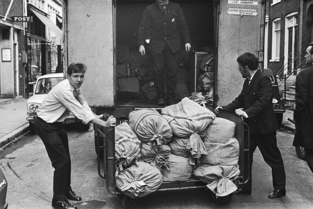 Eine Gruppe von Männern entlädt Taschen von einem geparkten Lkw an der Seite einer Straße, mit einem vorbeifahrenden Auto und Gebäuden mit Fenstern, Treppen mit Geländern und Texttafeln im Hintergrund, alles in Schwarz-Weiß dargestellt.