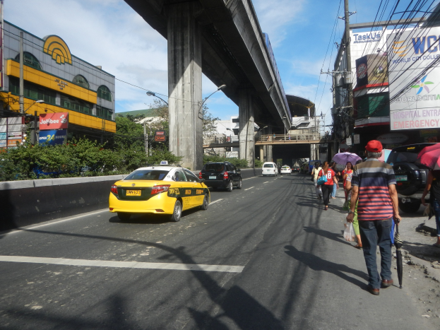 Ein gelbes Taxi fährt eine Stadtstraße entlang mit hohen Gebäuden, Fußgängern mit Regenschirmen auf dem Gehweg, einer Brücke im Hintergrund und bewölktem Himmel mit Strommasten und -drähten.