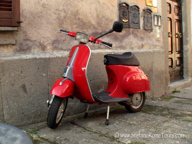 Ein rotes Rollerfahrrad steht vor einem Haus mit einer Tür und Fenstern auf einer Straße mit sichtbarem Text.