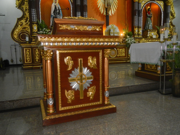 Ein Altar in einer Kirche mit einem Kreuz, Statuen, Blumensträußen, Kerzen, einem Tisch mit einer Decke und anderen Gegenständen, wobei die Wände mit Lichtern geschmückt sind.