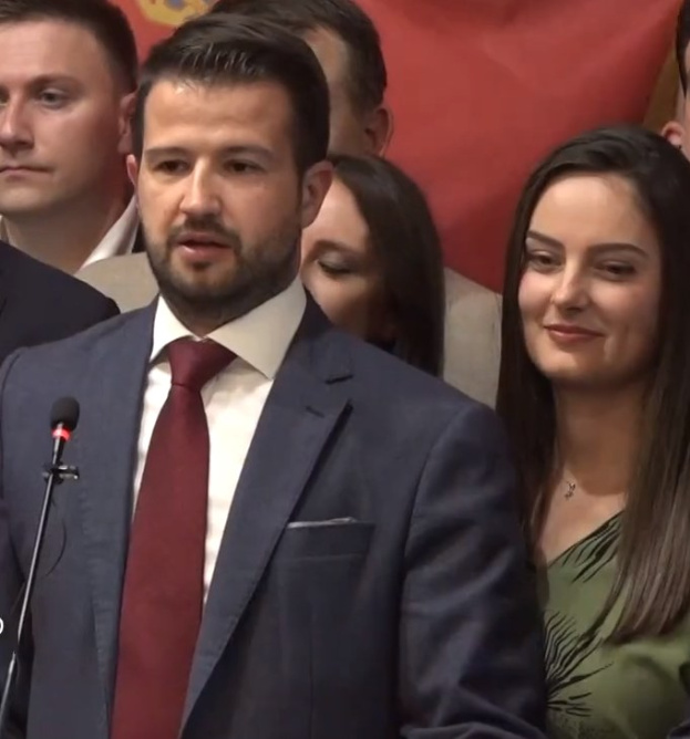 Ein Mann im Anzug und Krawatte steht an einem Rednerpult mit einem Mikrofon und spricht vor einem Publikum mit einem Banner im Hintergrund, wahrscheinlich wegen eines Regierungverbots für soziale Medien.