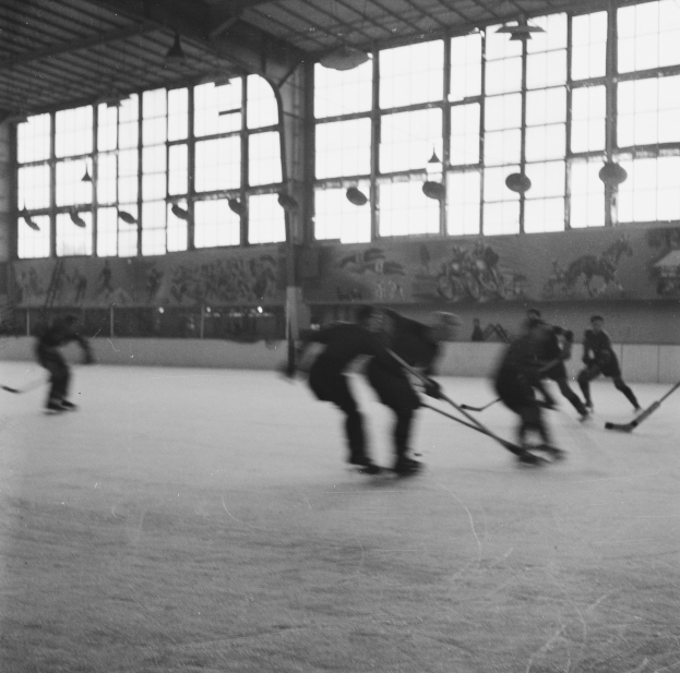 Schwarzes und weißes Bild einer Gruppe von Menschen, die Hockey auf einem Eisplatz spielen, mit einer bemalten Wand und Glasfenstern im Hintergrund.