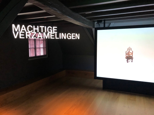 Installationsansicht der Ausstellung 'Machtige Verzamelingen' im Museum of Modern Art Berlin mit einem großen Bildschirm mit Text auf der rechten Seite, einem Fenster auf der linken Seite und sichtbarer Decke.