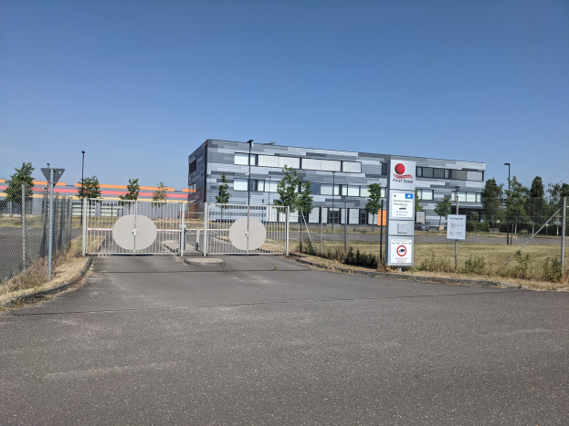 Großes industrielles Gebäude mit der Bezeichnung "First Solar" mit vielen Fenstern, umgeben von einem Metallzaun, umgeben von Bäumen, Pflanzen, Gras, Schildern, Straßenmasten und einem Weg unter einem bewölktem Himmel.