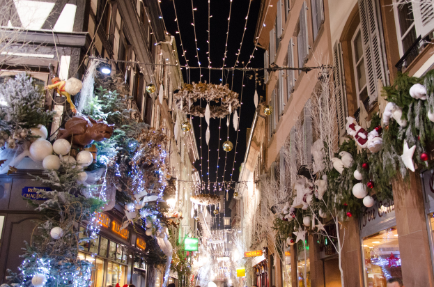 Ein lebendiger Weihnachtsmarkt in Straßburg, Frankreich, mit Menschen, die zwischen Gebäuden spazieren, die mit Weihnachtsbäumen, Lichtern und anderen festlichen Dekorationen geschmückt sind, unter einem sichtbaren Himmel.