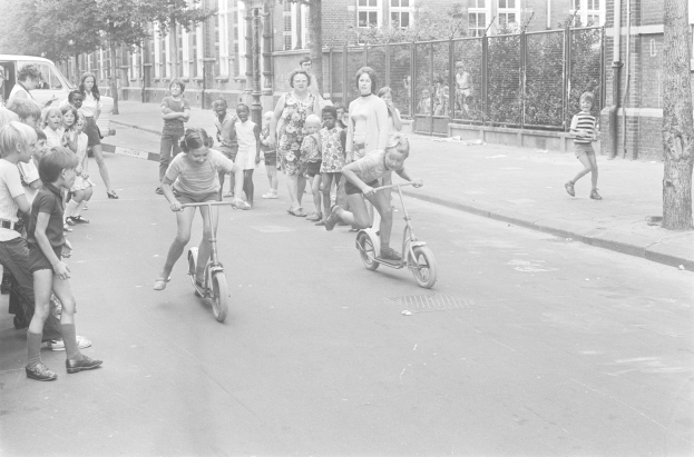 Schwarzes und weißes Bild von Kindern, die auf Roller die Straße entlangfahren, mit einem geparkten Auto auf der linken Seite, Gebäuden, Bäumen, Pfählen und einem Zaun im Hintergrund.