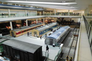 Ein großer Museumsraum mit verschiedenen Zügen in unterschiedlichen Größen und Farben, mit Menschen, die vom Boden, Geländern und Säulen unter Deckenlampen beobachten.