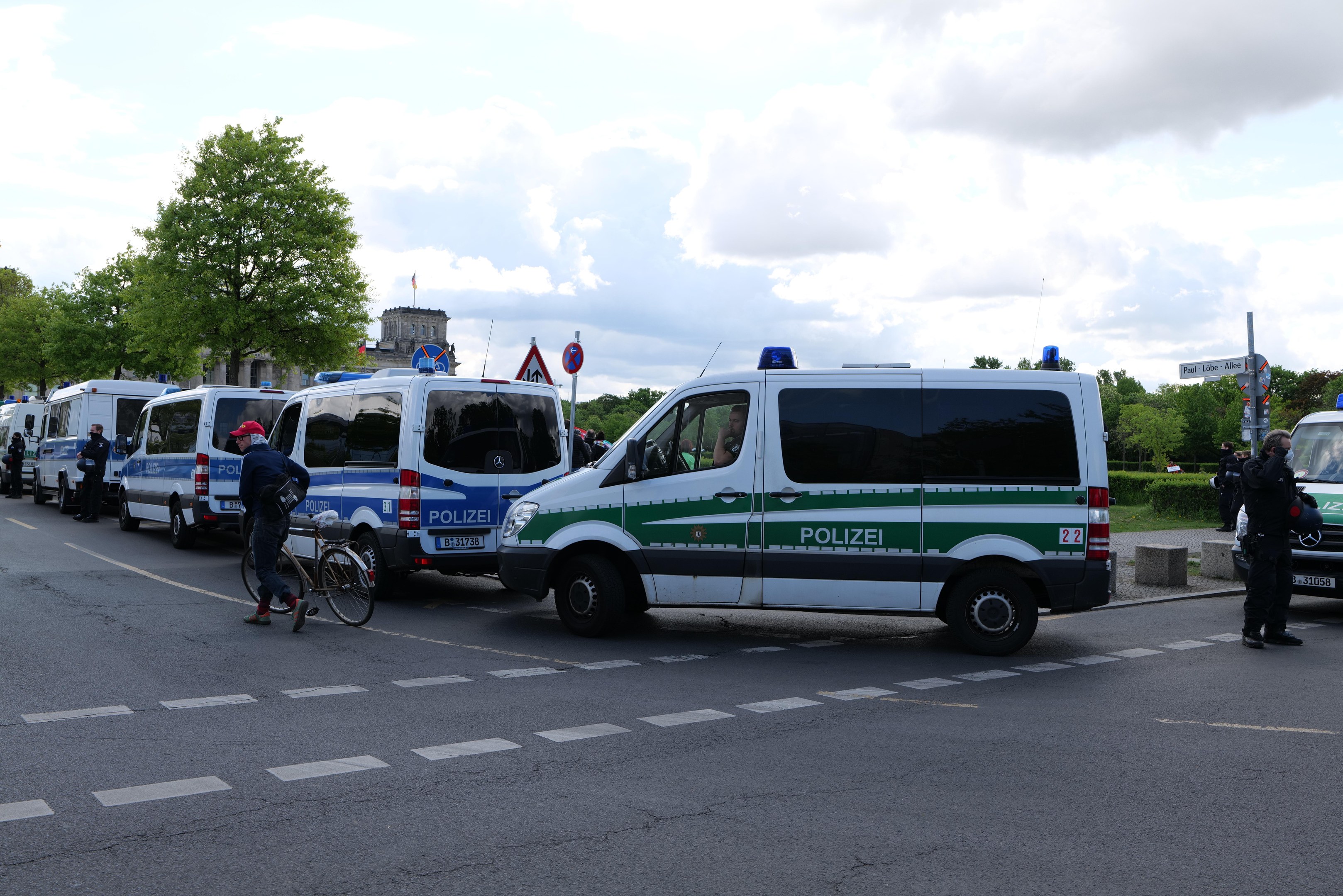 Polizeifahrzeuge parken entlang einer Straße mit einem Radfahrer im Vordergrund, Bäumen, Strommasten, Schildern, Gebäuden und einem bewölkten Himmel im Hintergrund.