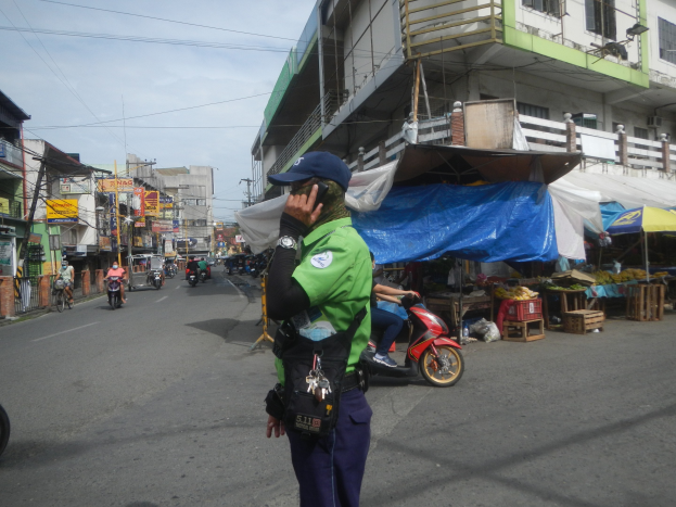Polizist in grüner und blauer Uniform mit blauer Mütze auf einer Straße, der mit einem Handy spricht, mit Fahrzeugen, Marktständen und einem Gebäude mit Fenstern im Hintergrund unter einem bewölkten Himmel.
