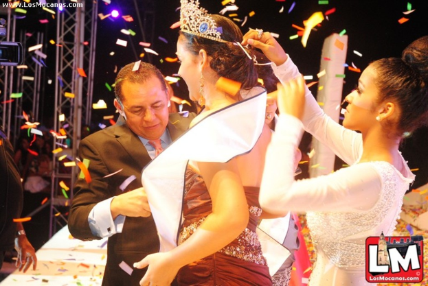 Ein Mädchen auf der Bühne bei einer Miss World Show mit einer Krone, mit einem anderen Mädchen, das sie von hinten anpasst, und einem Mann in einem schwarzen Anzug, der vor ihr die Hand schüttelt, vor einem dunklen Hintergrund mit Metallrahmen.