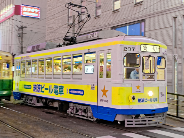 Eine gelbe und blaue Straßenbahn auf einer nächtlichen Stadtstraße mit Menschen darin, Gebäuden mit Fenstern, Strommasten mit Kabeln und Schildern im Hintergrund.